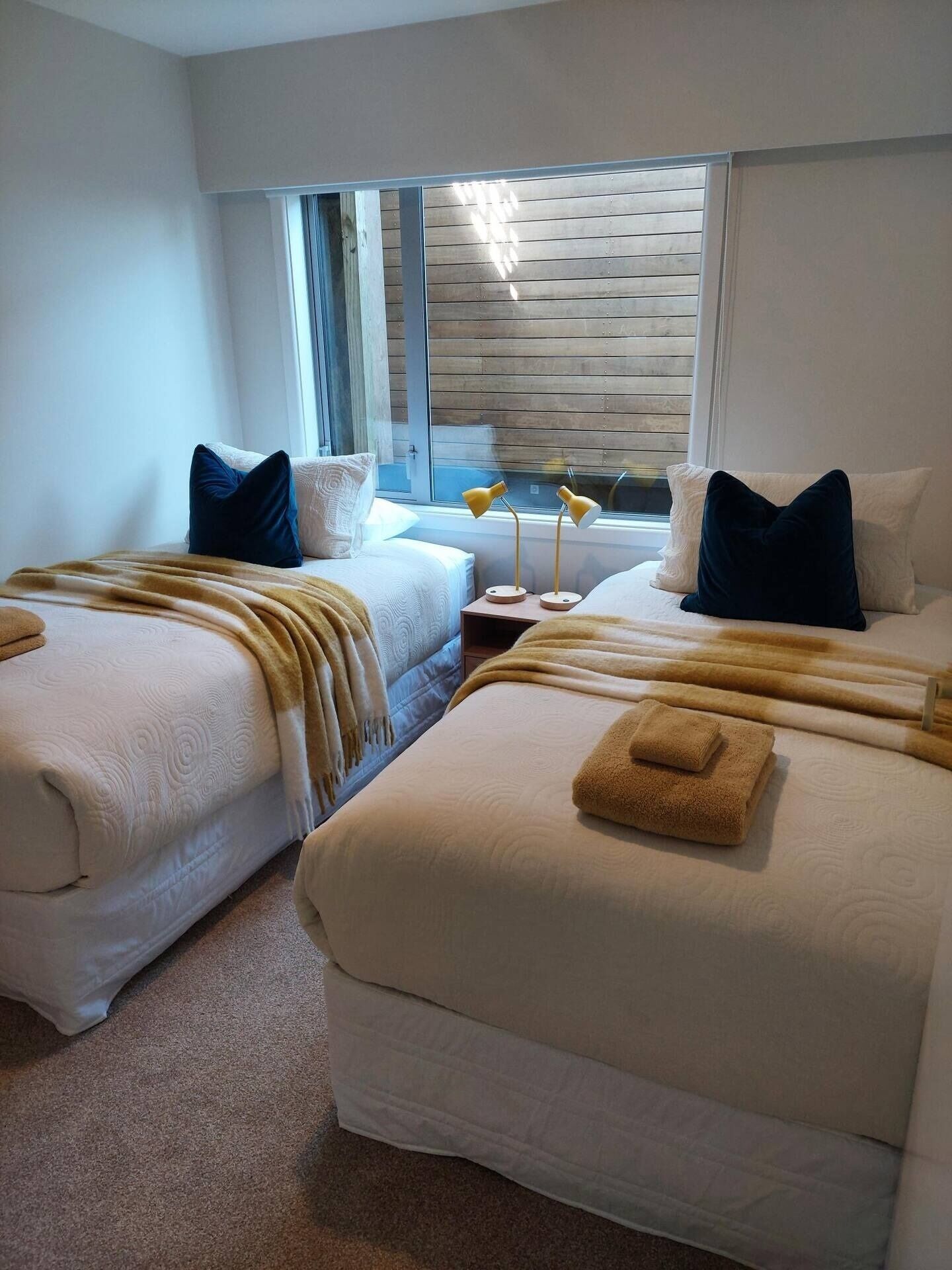 A hotel room with two beds, a window with wooden blinds, a bedside table with a lamp, and a view of the outside.