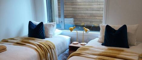 A hotel room with two beds, a window with wooden blinds, a bedside table with a lamp, and a view of the outside.