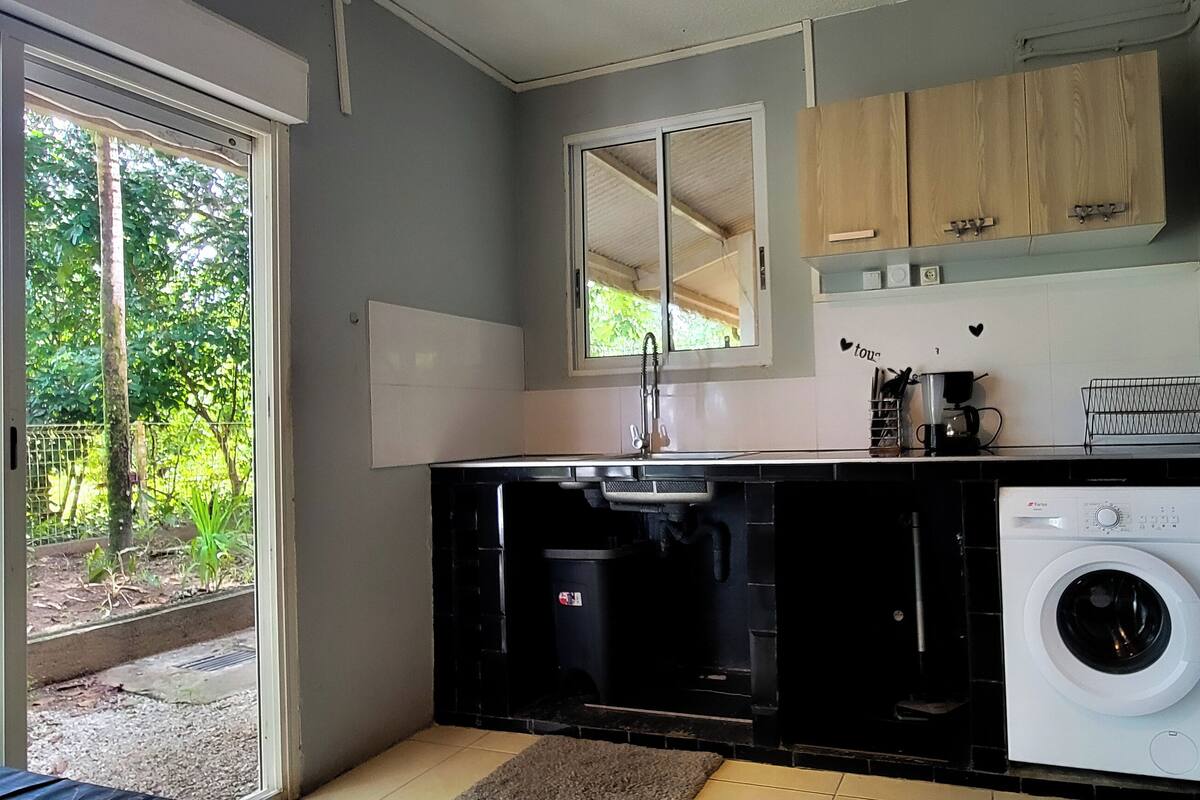 A compact kitchen with a washing machine, a sink, and a window overlooking greenery.