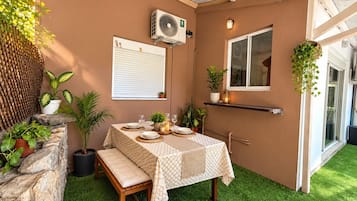 Un área de comedor al aire libre con una mesa dispuesta para dos personas, rodeada de plantas en macetas y un muro de piedra.