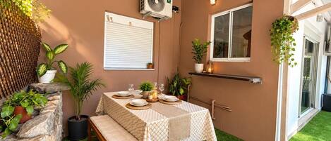 Un área de comedor al aire libre con una mesa dispuesta para dos personas, rodeada de plantas en macetas y un muro de piedra.