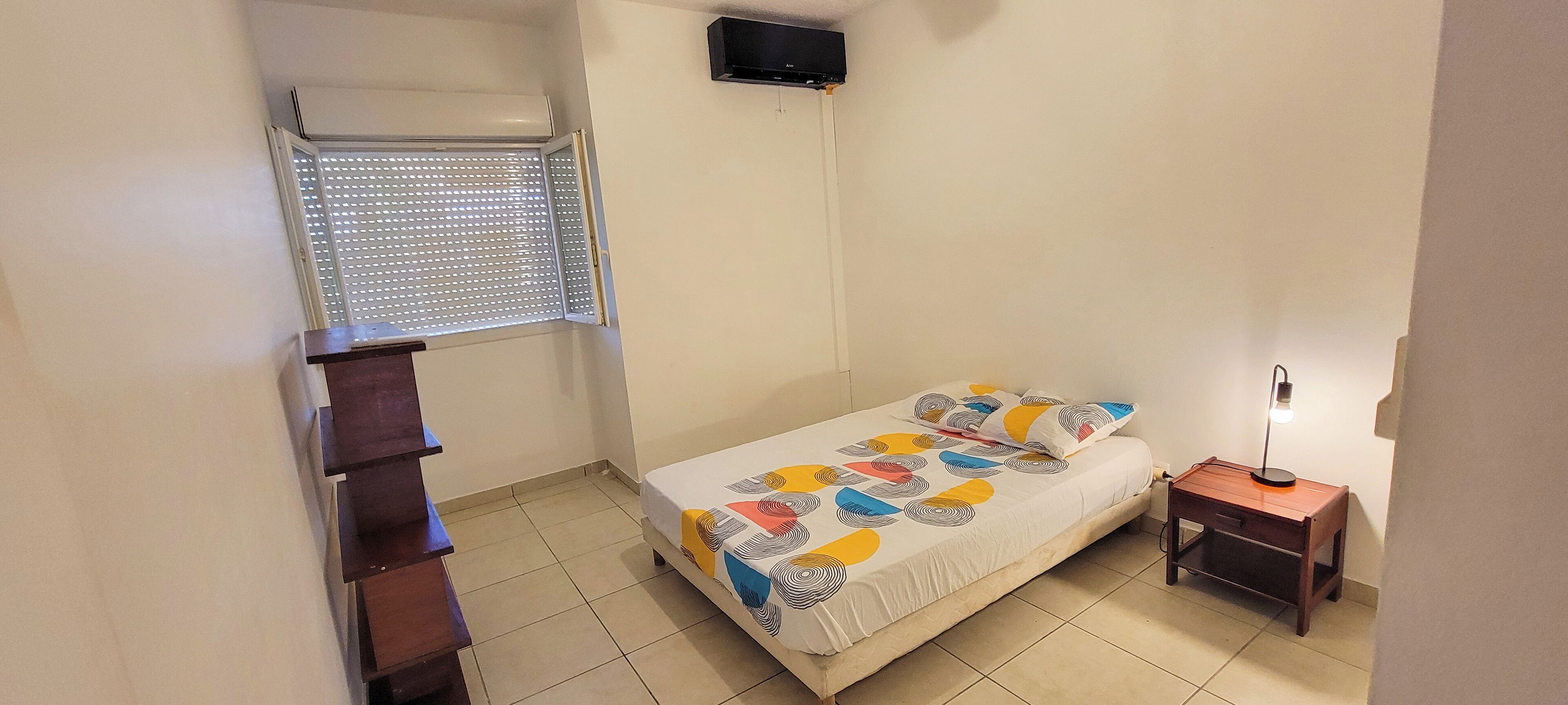 Um quarto pequeno e bem iluminado, com uma cama, mesa de cabeceira, janela com persianas, ventilador de teto e uma escada.