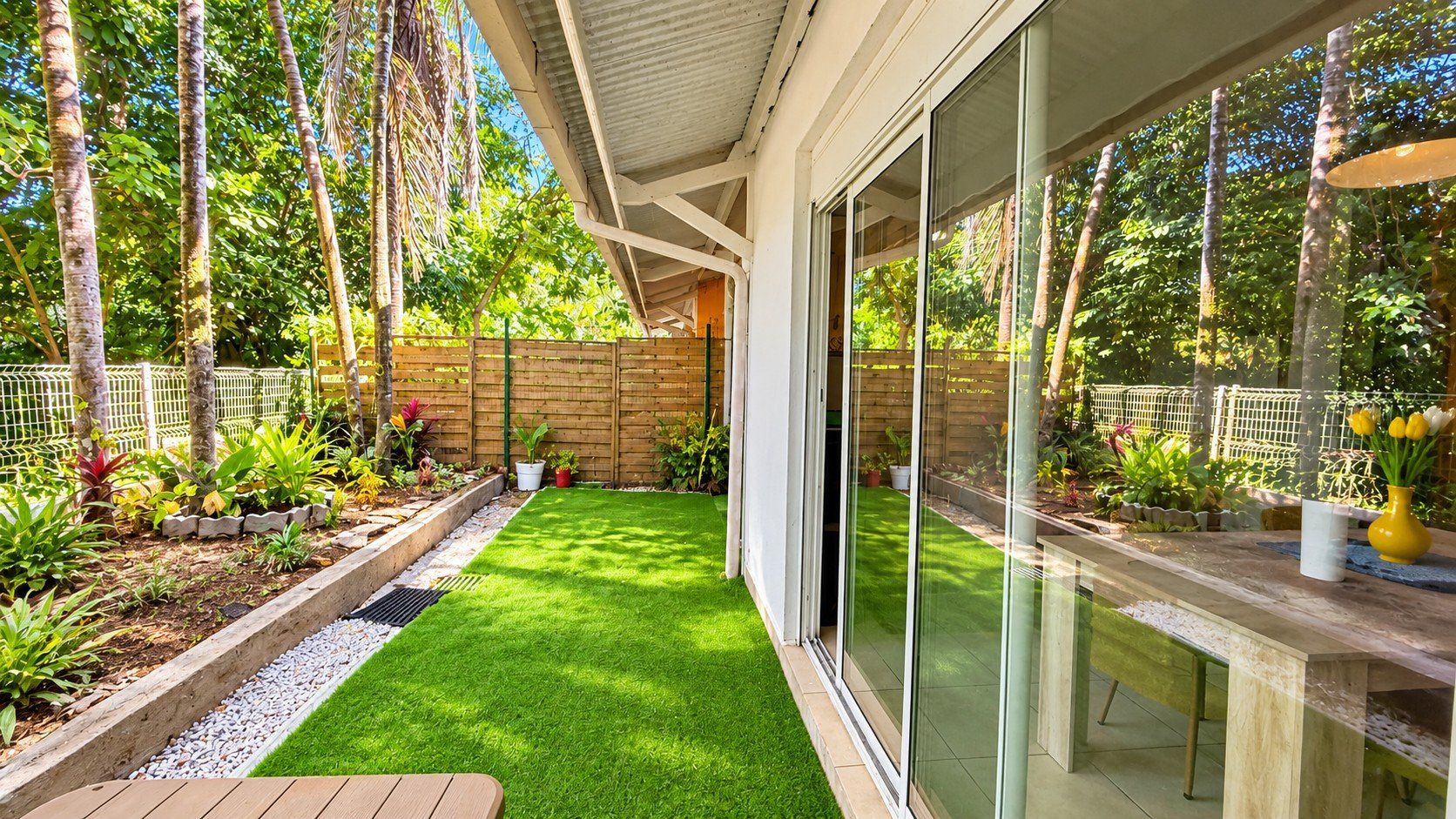 Une maison moderne dotée d’une porte vitrée donnant sur un jardin bien entretenu, avec une pelouse artificielle et diverses plantes.