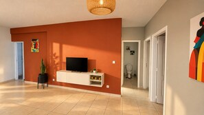 Un salon moderne avec un téléviseur à écran plat posé sur un meuble blanc, une plante en pot et une œuvre d’art colorée accrochée au mur.