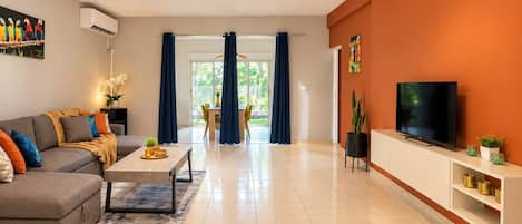 A modern living room with a sofa, coffee table, TV, and a dining area with chairs and curtains.
