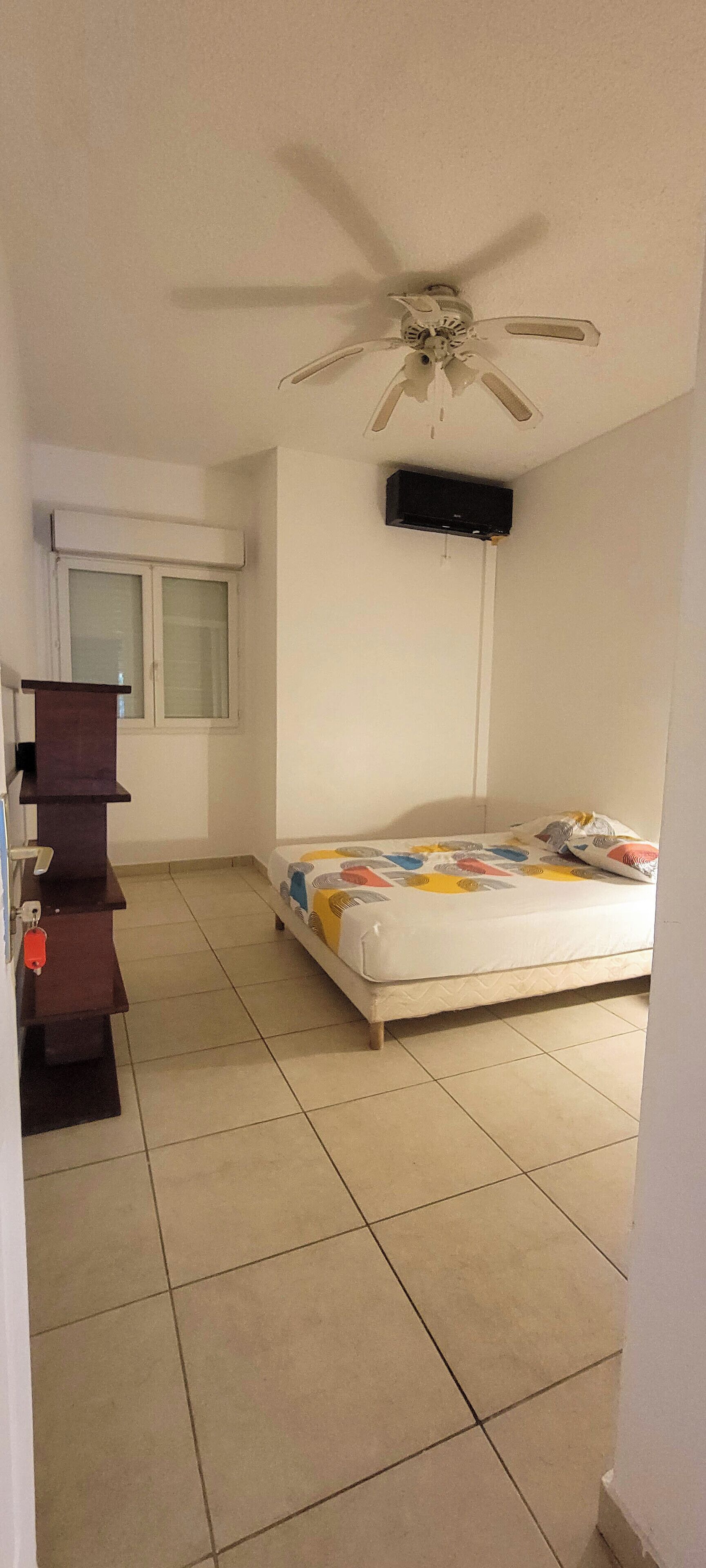 A bedroom with a bed, a ceiling fan, a window, and a TV mounted on the wall.