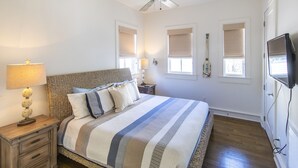 A bedroom with a bed, a nightstand, a lamp, a ceiling fan, and a TV mounted on the wall. - Starfish Carriage House (Rosemary Beach)
