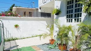 Un immeuble résidentiel doté d’un balcon, de plantes en pot et d’un petit espace jardin aménagé avec du gazon synthétique et du gravier.