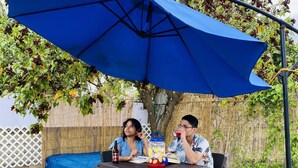 Deux personnes sont assises à une table à l’extérieur, sous un parasol bleu. Une bouteille et un récipient sont posés sur la table.