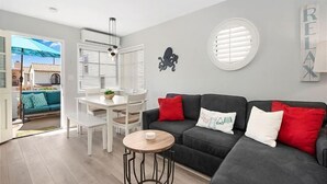 A modern living room with a grey sofa, a round wooden coffee table, and a white dining table with chairs. There is a window with blinds, a ceiling fan, and a wall-mounted decoration.