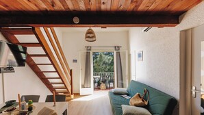 En hyggelig stue med trælofter, en grøn sofa, et spisebord med bøger og et lys, samt en trappe op til en balkon med udendørs møbler.
