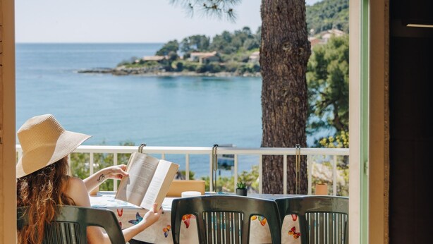 Une personne est assise à une table, devant un livre, et peut admirer la mer et un arbre.