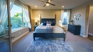 Une chambre à coucher comprenant un lit, un ventilateur de plafond, une fenêtre avec des rideaux, une table de chevet et une commode.