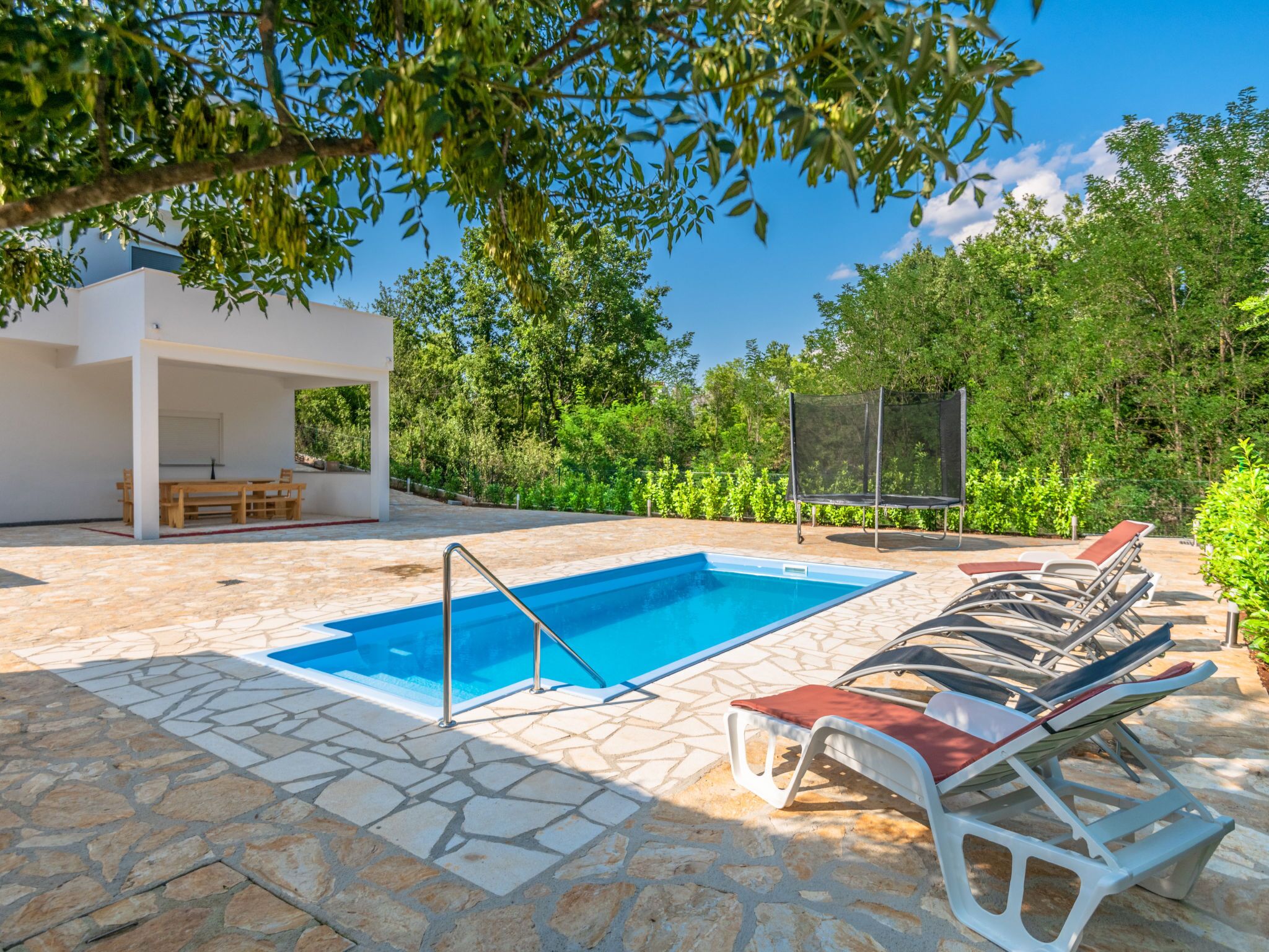 Una piscina con terraza de piedra, sillas de descanso y un área de estar techada.
