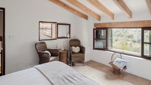Un dormitorio con cama, dos sillones de mimbre, una mesita, un espejo y una ventana con vistas al exterior.