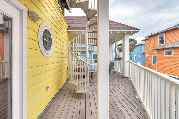 A wooden deck with a white railing, a yellow building with a window, and a blue building in the background.