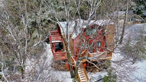 Une cabane rouge avec un escalier en bois et un petit porche, entourée d’arbres couverts de neige.