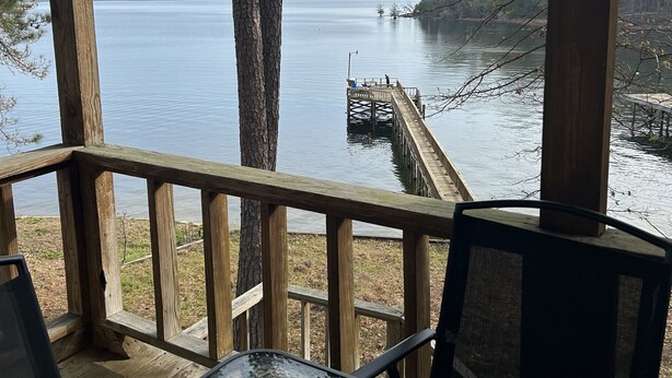 Una terraza de madera con una mesa y sillas de vidrio, con vista a un lago tranquilo y un muelle que se adentra en el agua.