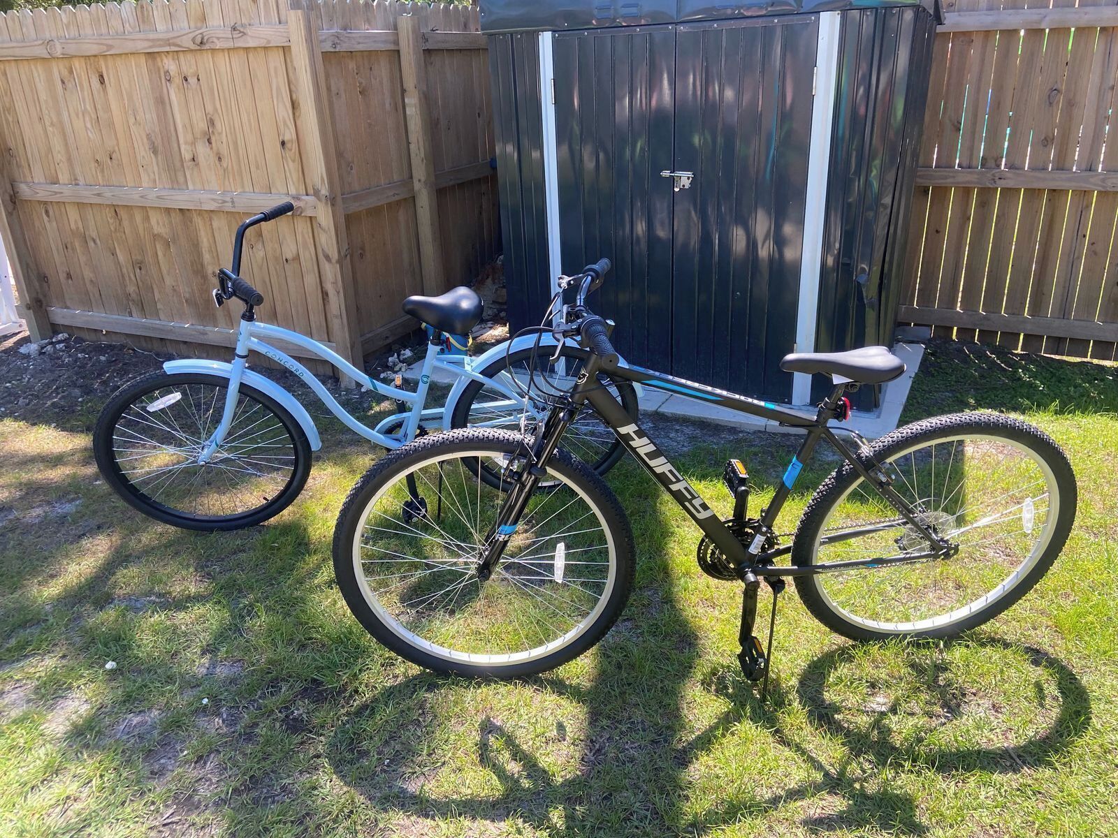 Twee fietsen staan op het gras, met een houten hek en een tuinhuis op de achtergrond.