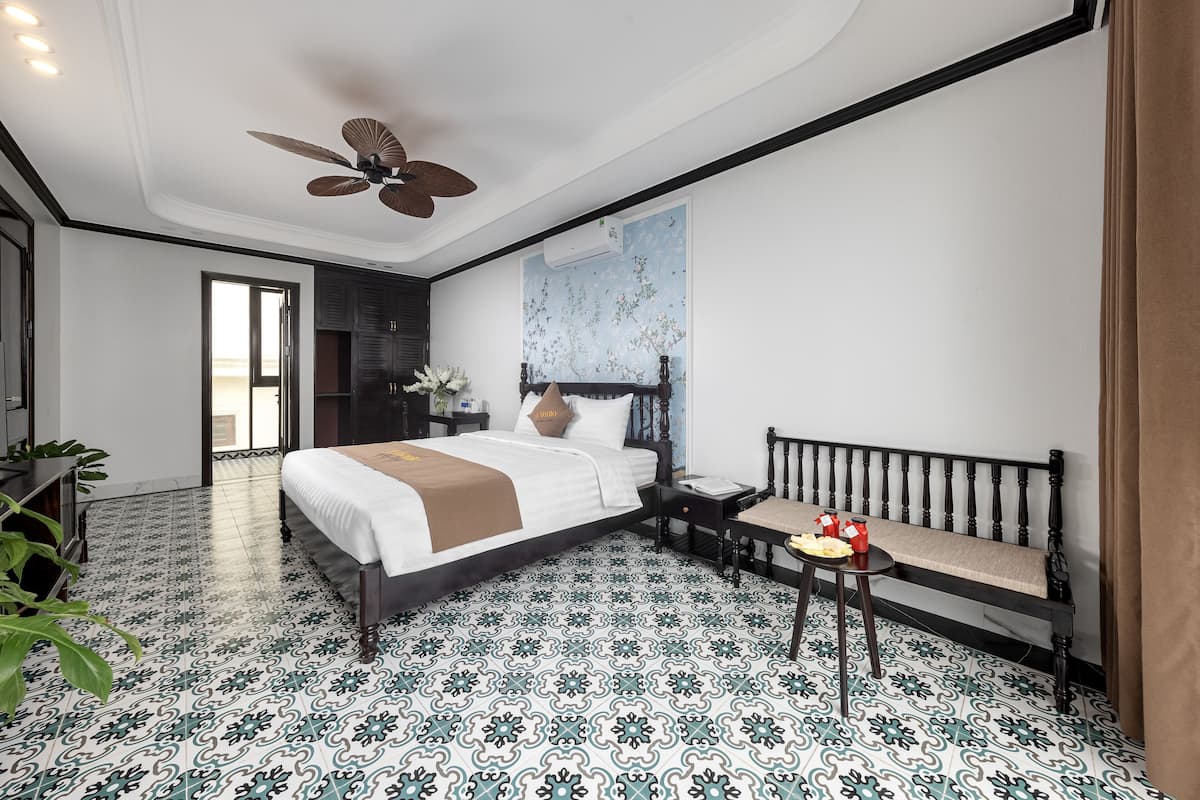 A hotel room with a bed, a bench, a small table with snacks, a ceiling fan, and patterned floor tiles.