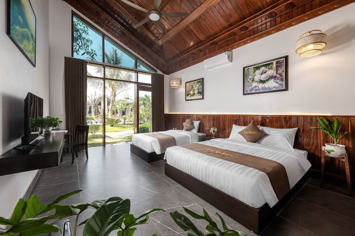 A modern hotel room with two beds, a TV, a dining area, and large windows offering a view of the outdoors.