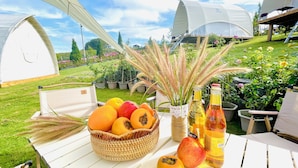 A white table with a basket of fruits, a bottle, and a chair in a garden with tents.