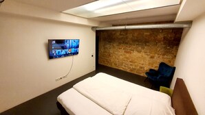 Habitación de hotel con una cama, un televisor montado en la pared, un muro de ladrillo y una silla azul.