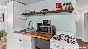 A compact kitchen with a built-in microwave, stove, and sink. There is a wooden shelf above the stove holding spices and a red container. - Borealis Barnhouse - Slumber 7 (Anchorage)