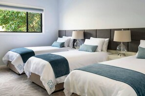 Ein Hotelzimmer mit drei Betten, jedes mit einer weiß-blauen Tagesdecke, und einem grauen Kopfteil. Zwei Nachttische mit klaren Lampenschirmen sind vorhanden.