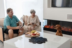 A living room with a fireplace, a coffee table with snacks, and two people seated on a sofa. - Vue One Nelson Bay Waterfront Luxury (Nelson Bay)