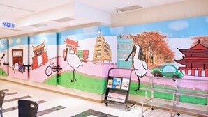 Eine Wand mit farbenfrohen Wandgemälden verschiedener Gebäude und einem Storch. Stühle und ein Wagen mit Zeitschriften stehen auf dem Boden.