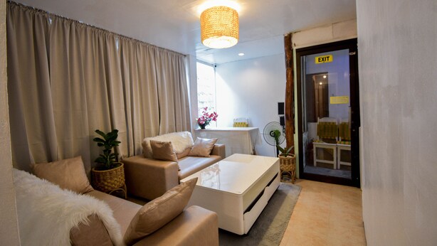 A modern living room with a sofa, a white coffee table, and a lamp. There is a door with an "EXIT" sign and a window with curtains. - Treasure Isle Backpacker's Hostel (Boracay Island)