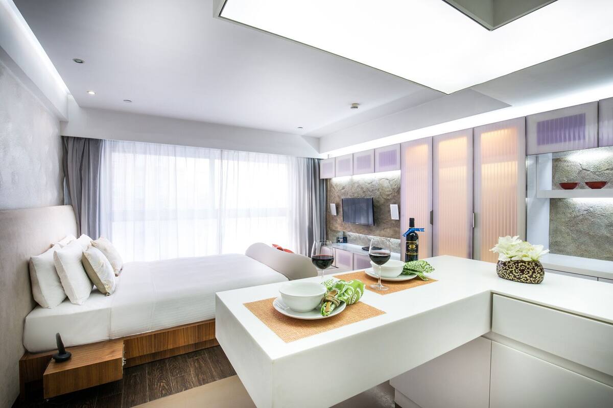 A modern hotel room with a bed, a dining area, a kitchen, and a TV mounted on the wall.