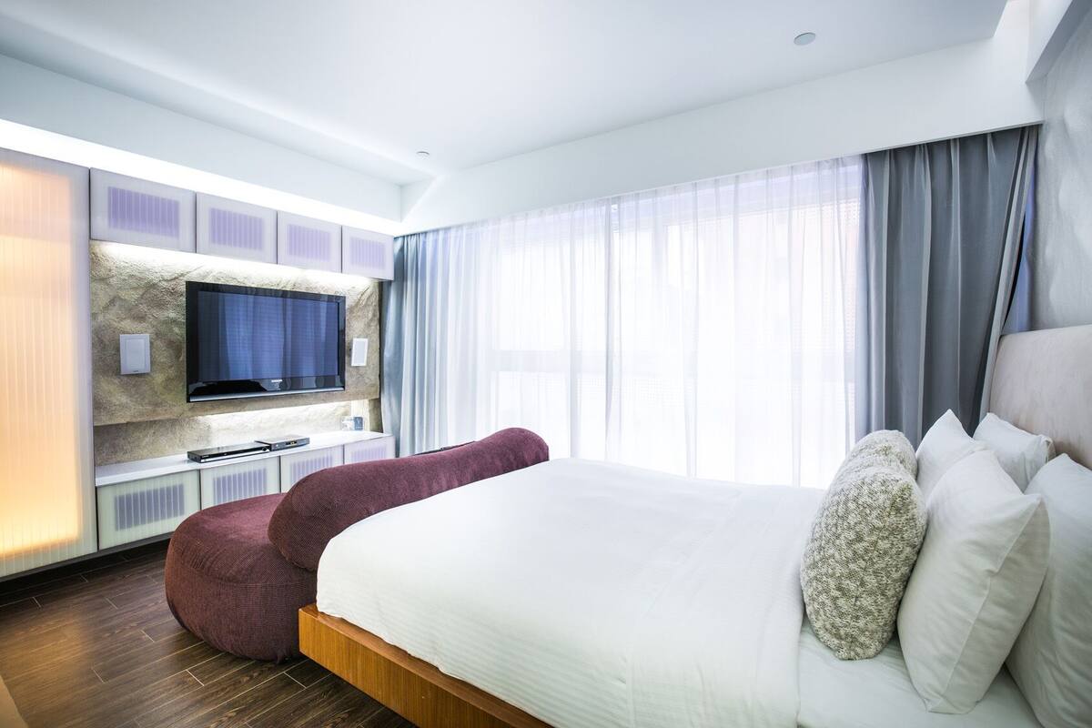 A modern hotel room with a large bed, a flat-screen TV mounted on a textured wall, a seating area with a red chair, and a large window with sheer curtains.