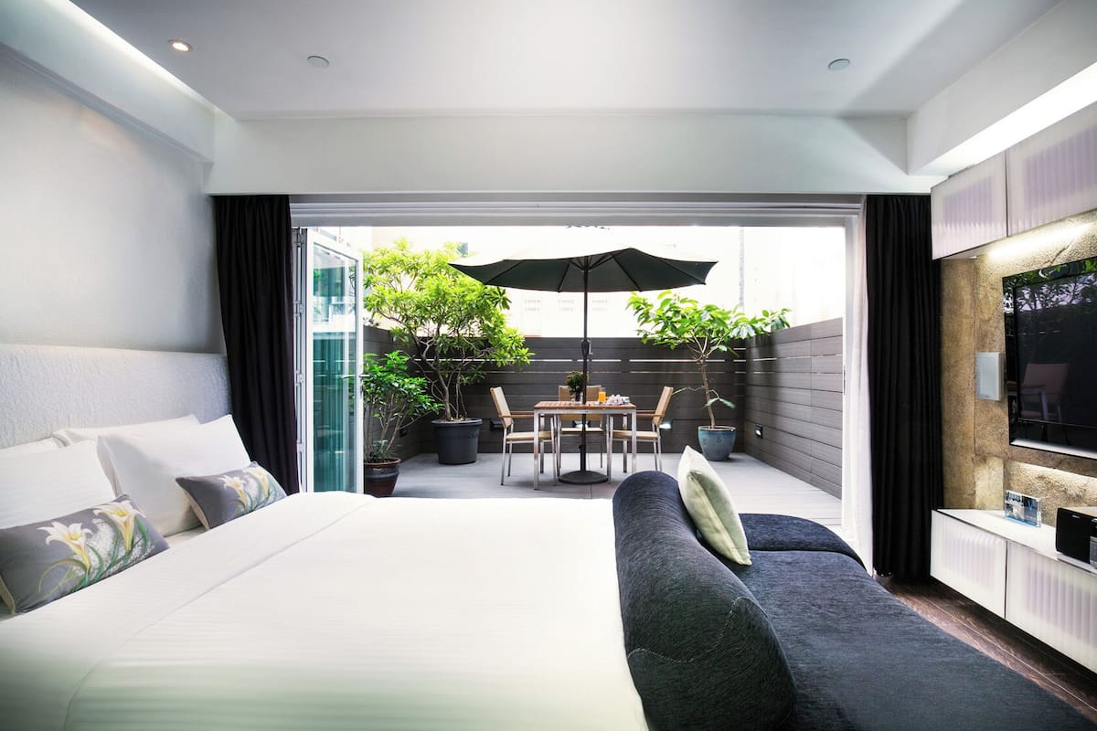 A modern hotel room with a large bed, a sofa, a dining area with an umbrella, and a TV.
