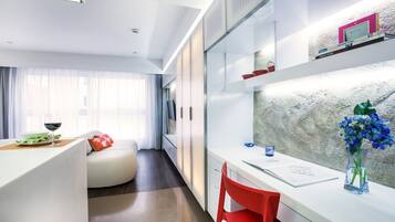 A modern hotel room with a dining area, a bed with a red pillow, a desk with a vase of flowers, and a view of the city through large windows.