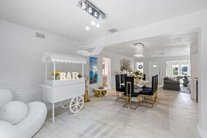 Un interior moderno con un carrito bar blanco, una zona de comedor con sillas negras y una mesa blanca, y una araña.