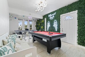 Una mesa de billar con un letrero de neón, una mesa de pool y una pared verde con follaje.