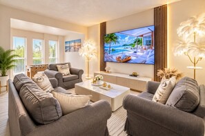 A modern living room with a large flat-screen TV, grey sofas, a coffee table, and a large window with blinds.