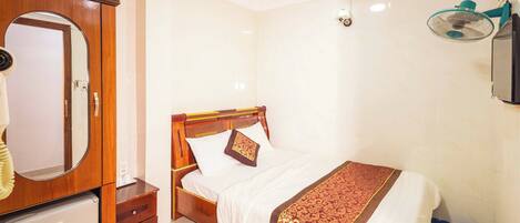 A bedroom with a wooden bed, a mirror, a small refrigerator, and a wall-mounted fan.