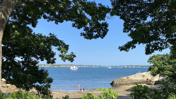 A coastal beach scene with clear skies, a sailboat, and people on the shore. - 5 minute walk to Beach, Stage Fort Park, and Gloucester Promenade! (Gloucester)