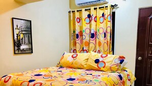 Une chambre à coucher avec un lit recouvert d’une couvre-lit à motifs floraux, une tête de lit en bois, un climatiseur fixé au mur, une porte et un tableau encadré accroché au mur.
