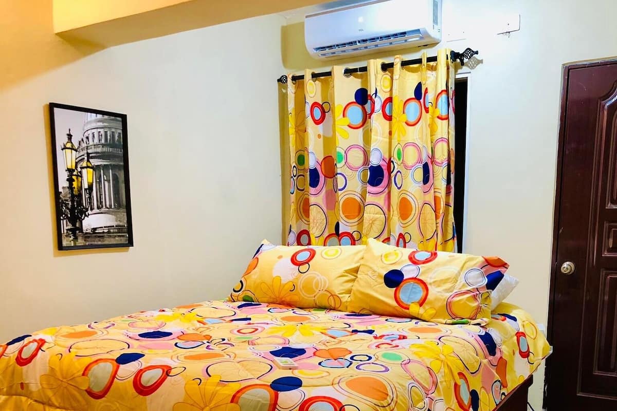 Ein Schlafzimmer mit einem Bett, das mit einer blumigen Tagesdecke bezogen ist, einem Holz-Kopfteil, einer Wand-Klimaanlage, einer Tür und einem gerahmten Bild an der Wand.