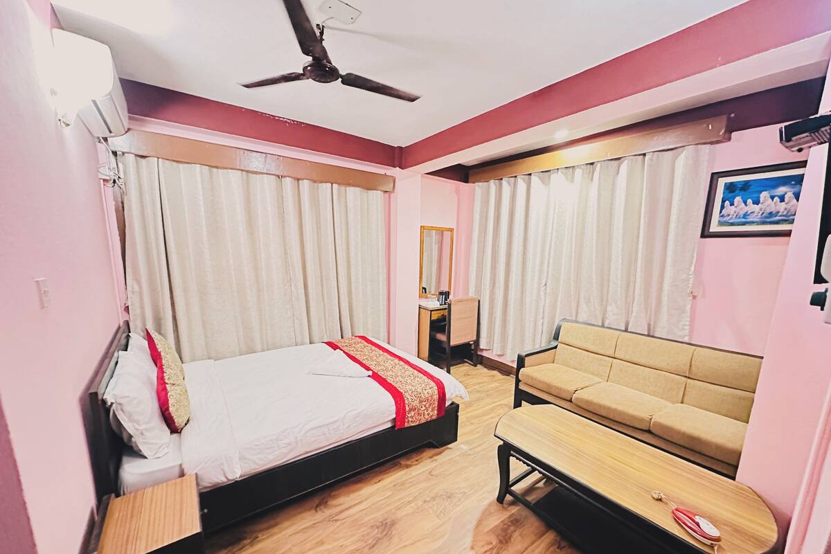 A hotel room with a bed, a sofa, a small table, a ceiling fan, and curtains.