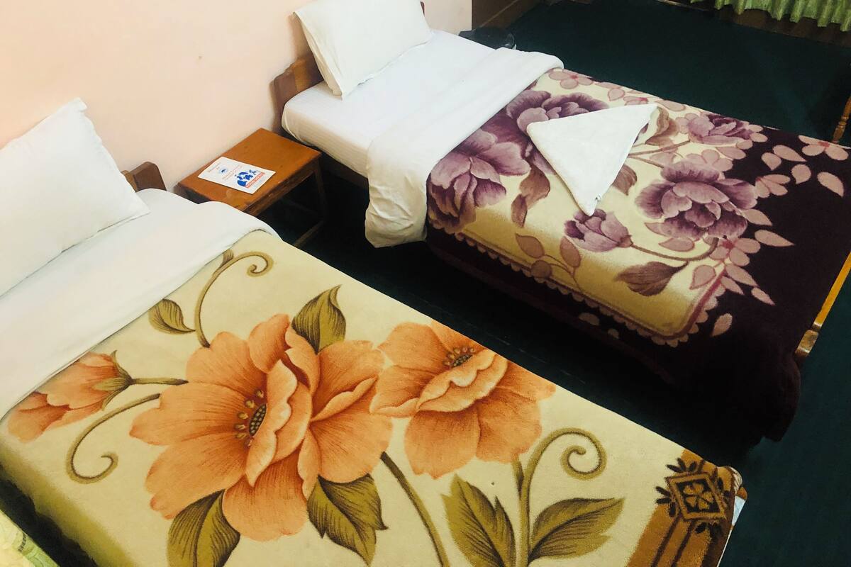 A hotel room with two beds, one with a floral bedspread and the other with a plain white bedspread. There is a small wooden nightstand with a lamp and a card on it.