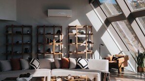 Amplia sala de estar con techo alto, candelabro y ventanas grandes que dejan entrar luz natural. Se observan varias estanterías de madera y un sofá con cojines.