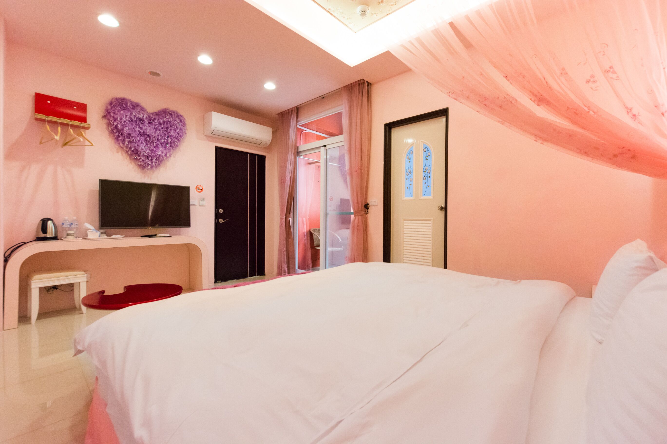 Een hotelkamer met een bed, een televisie, een bureau, een rode stoel, een paarse hart decoratie en een deur met een ruit.