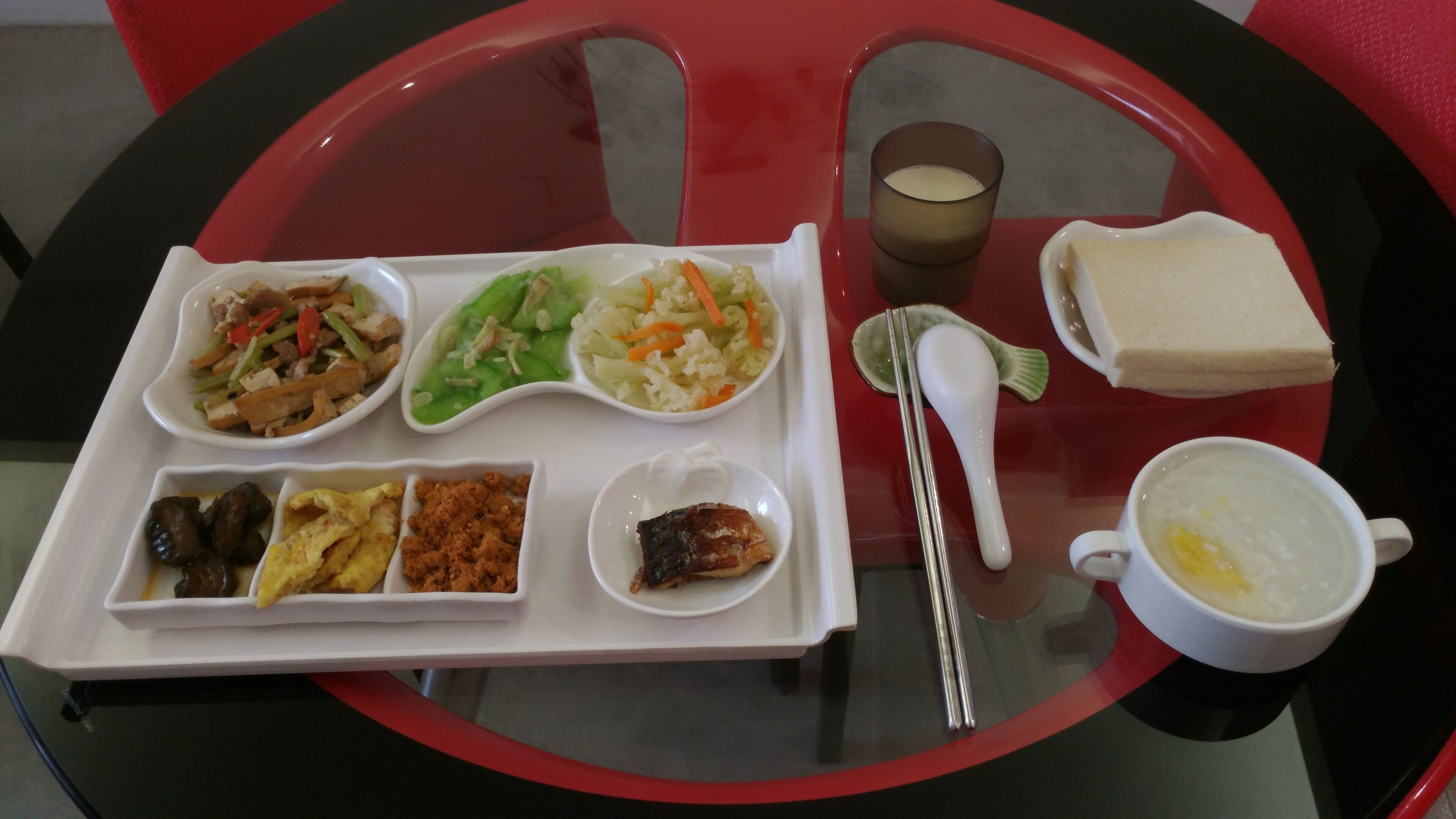 Un plateau contient divers plats, notamment des légumes sautés, une salade et un petit bol qui semble contenir un plat de viande. On y trouve également une tasse de soupe et un verre de lait.