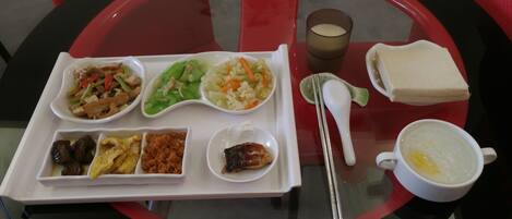 Un plateau contient divers plats, notamment des légumes sautés, une salade et un petit bol qui semble contenir un plat de viande. On y trouve également une tasse de soupe et un verre de lait.