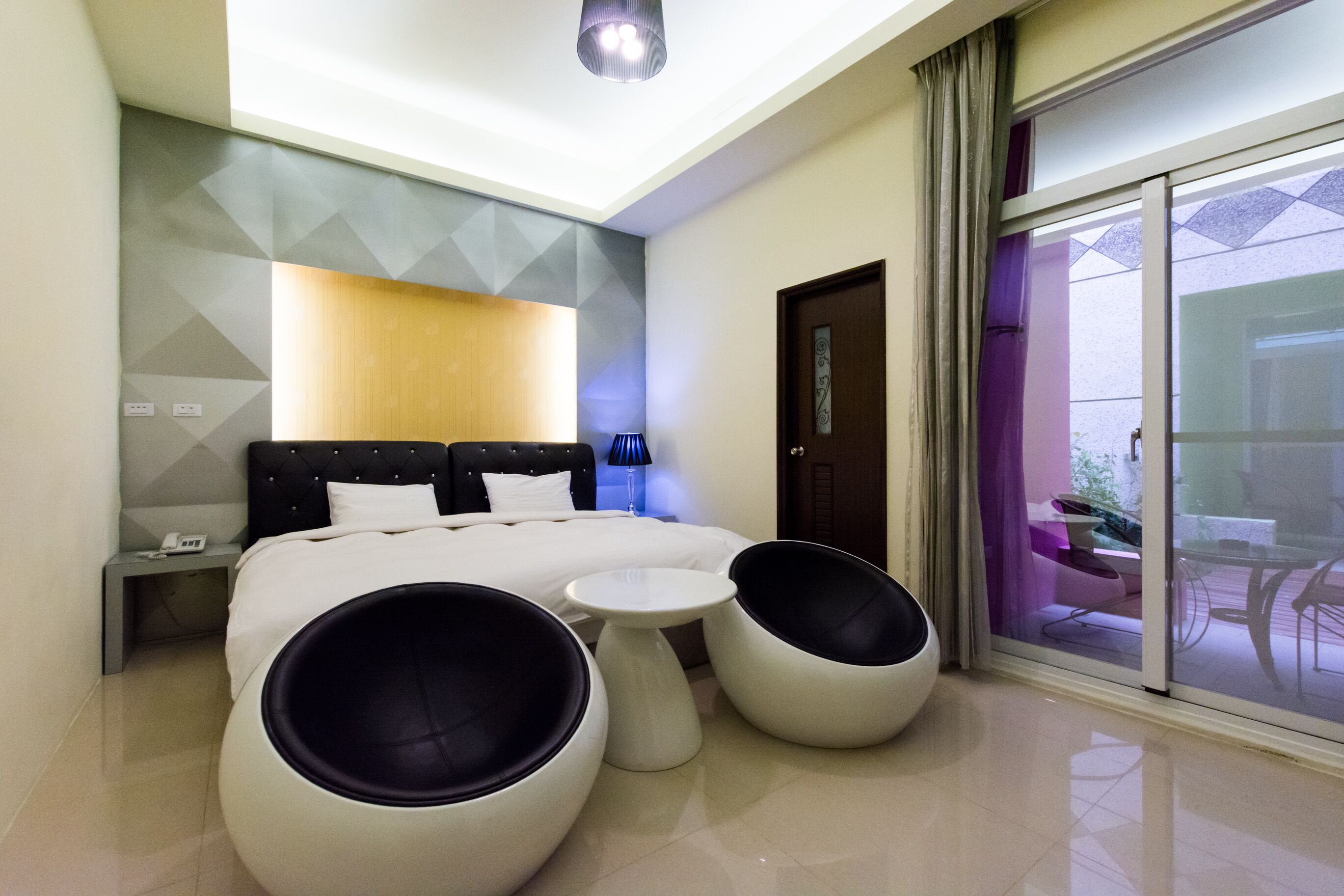 A modern hotel room with a large bed, two round seating areas, a small table, and a view through a sliding door to an outdoor area.